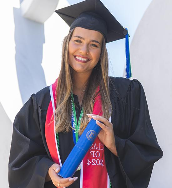 A UWF graduate smiling in cap and gown attire while holding a diploma tube.
